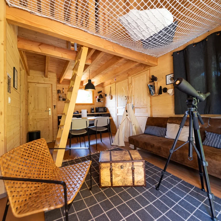 Salon en bois de la cabane Tribu avec canapé, kitchenette, télescope et filet suspendu en mezzanine.